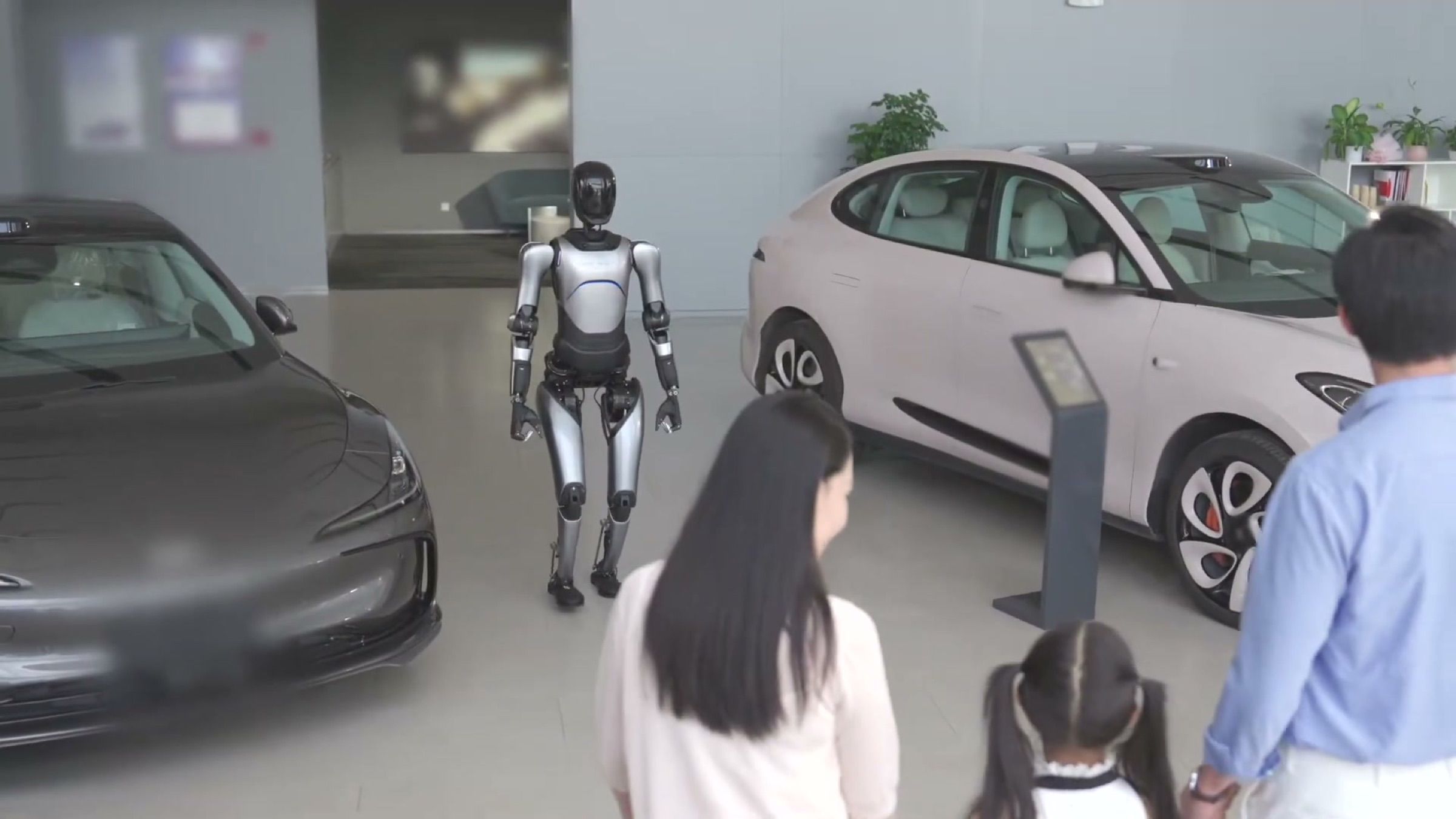 A humanoid robot walks between display cars to greet a family arriving at a car dealership.
