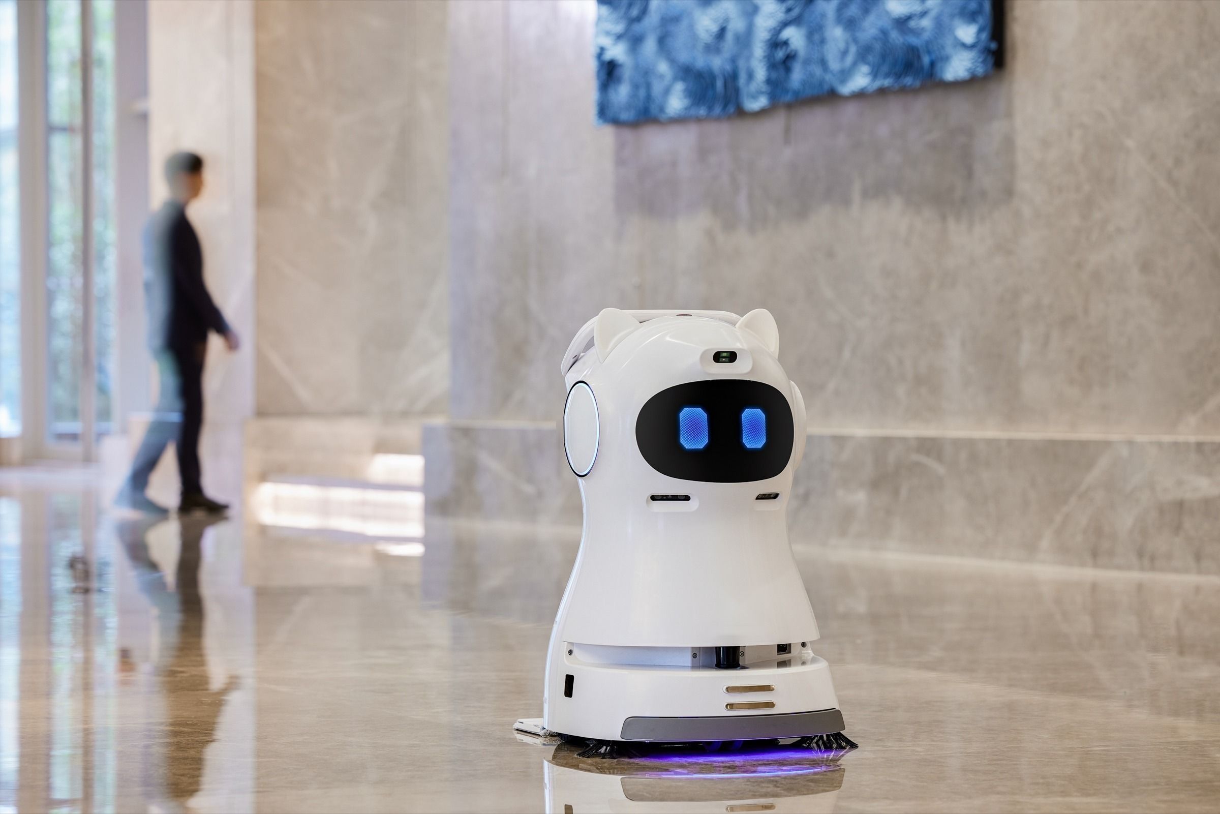 Autonomous cleaning robot working a polished lobby floor while a pedestrian walks past
