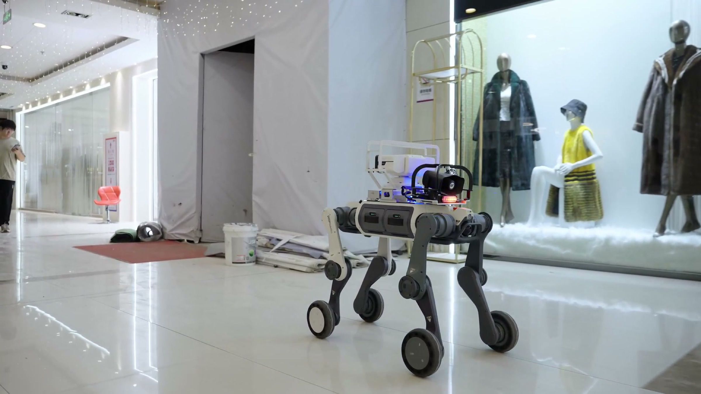 A wheeled-legged security robot with a camera and speaker patrols a quiet shopping centre concourse past shop windows and leftover renovation debris (frame at 0:31).
