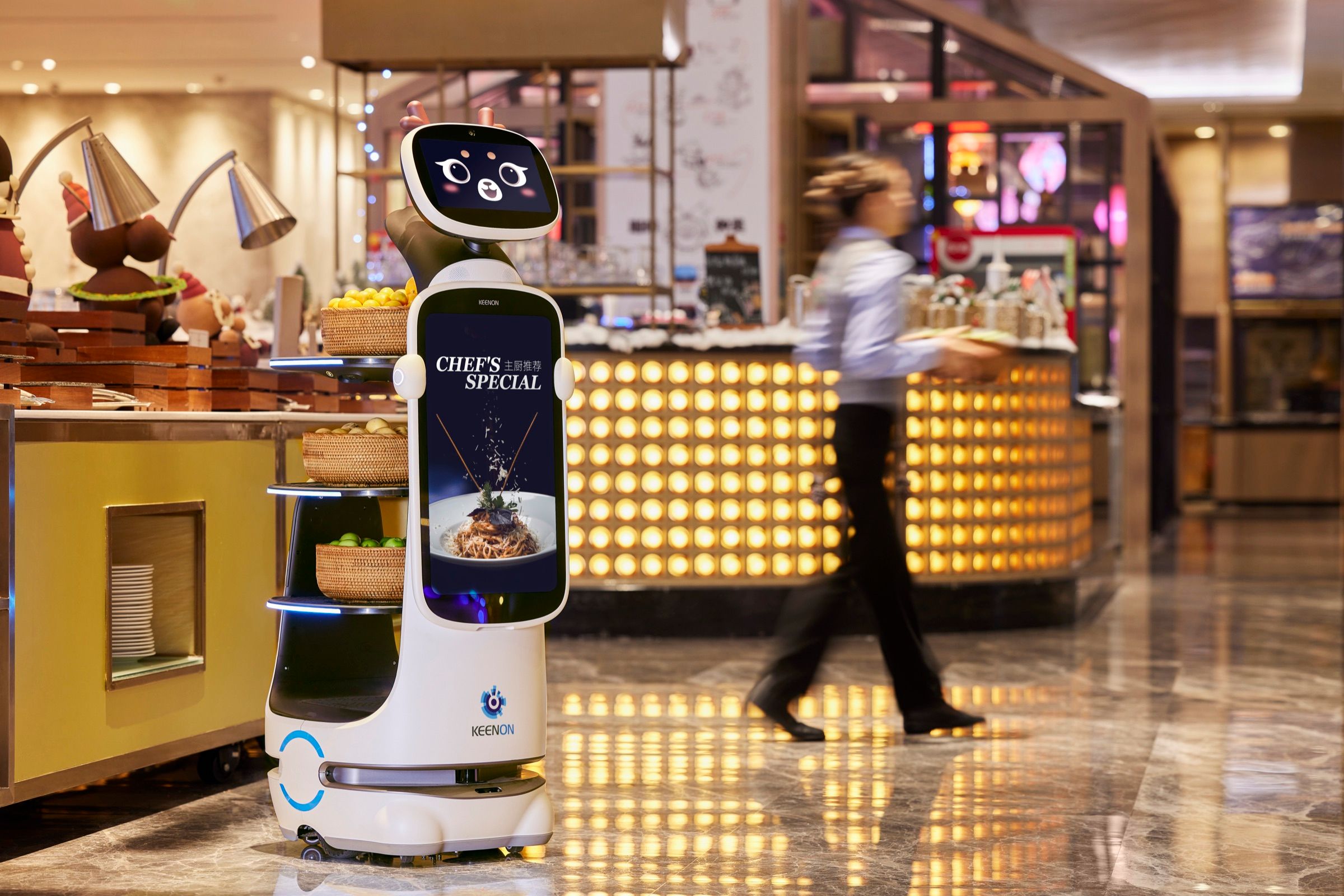 Service robot carrying a dish past a buffet counter as a staff member walks by in a hotel restaurant