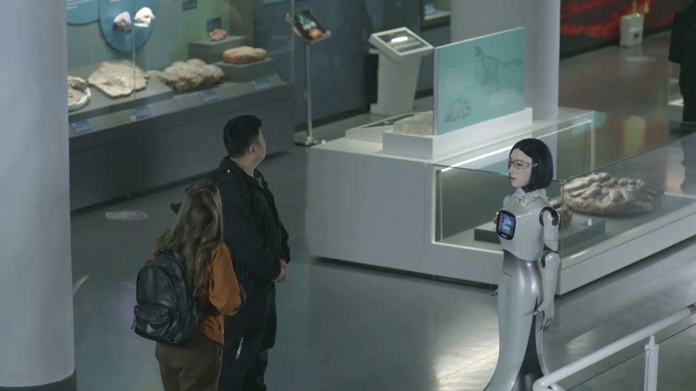 A humanoid guide robot talks two visitors through a fossil display in a museum gallery. Frame at 34.0s.