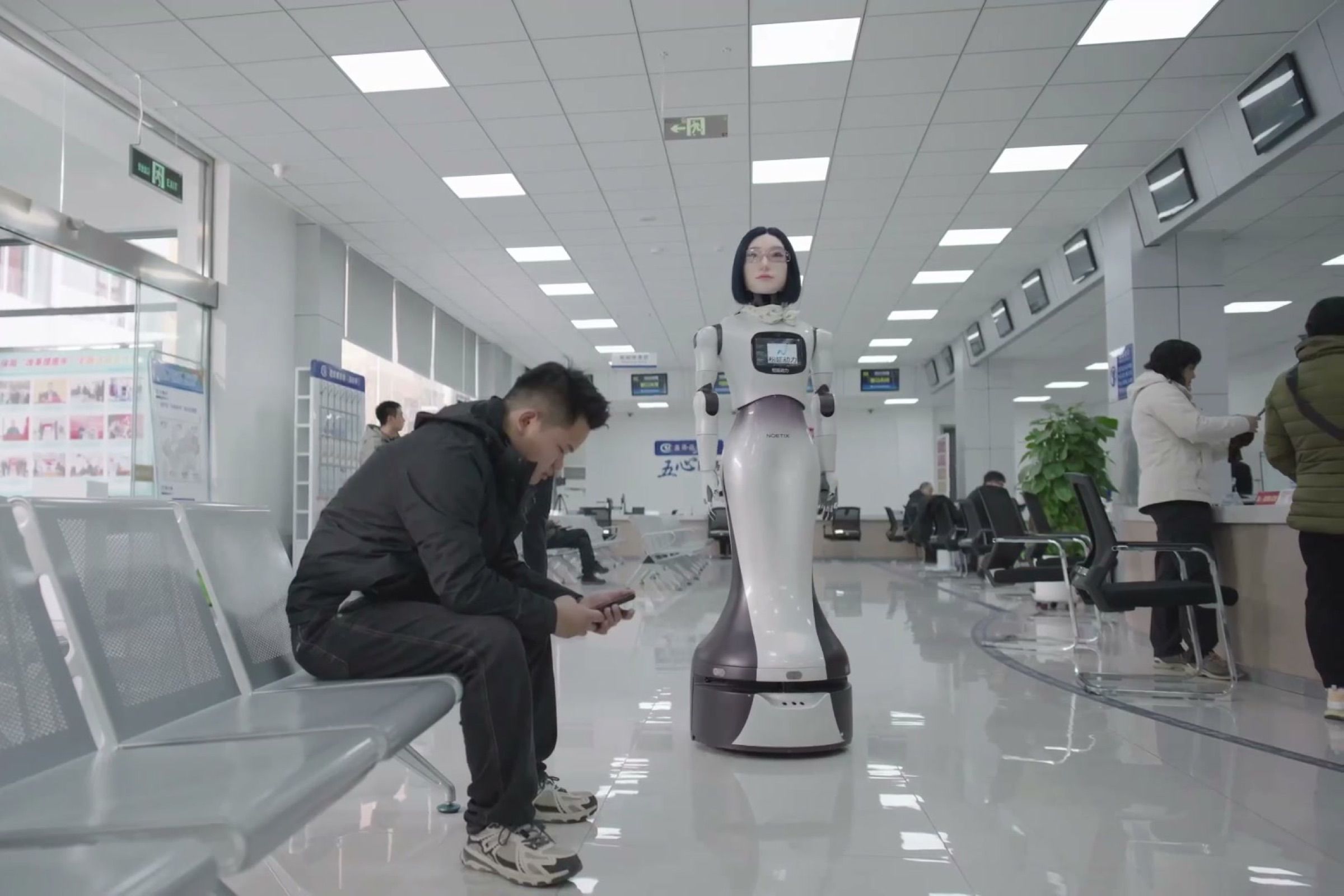 A wheeled humanoid robot checks on a visitor waiting in a public service hall, with service counters behind. Frame at 60.8s, cropped to remove a wall sign and an overlay.