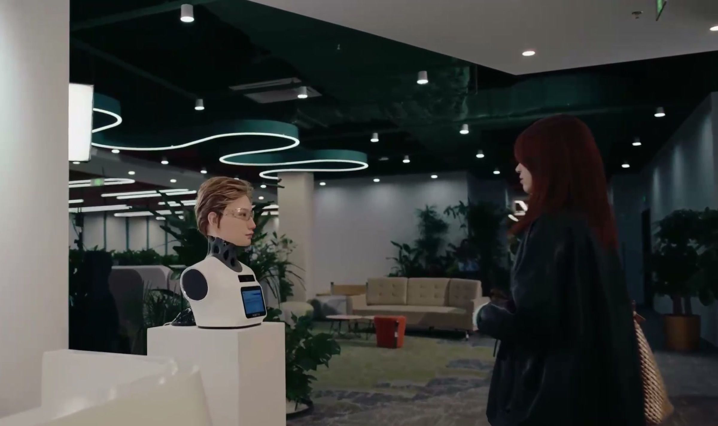 A lifelike desktop reception robot helps a visitor at an office front desk. Frame at 27.0s, cropped to remove a subtitle and a monitor logo.
