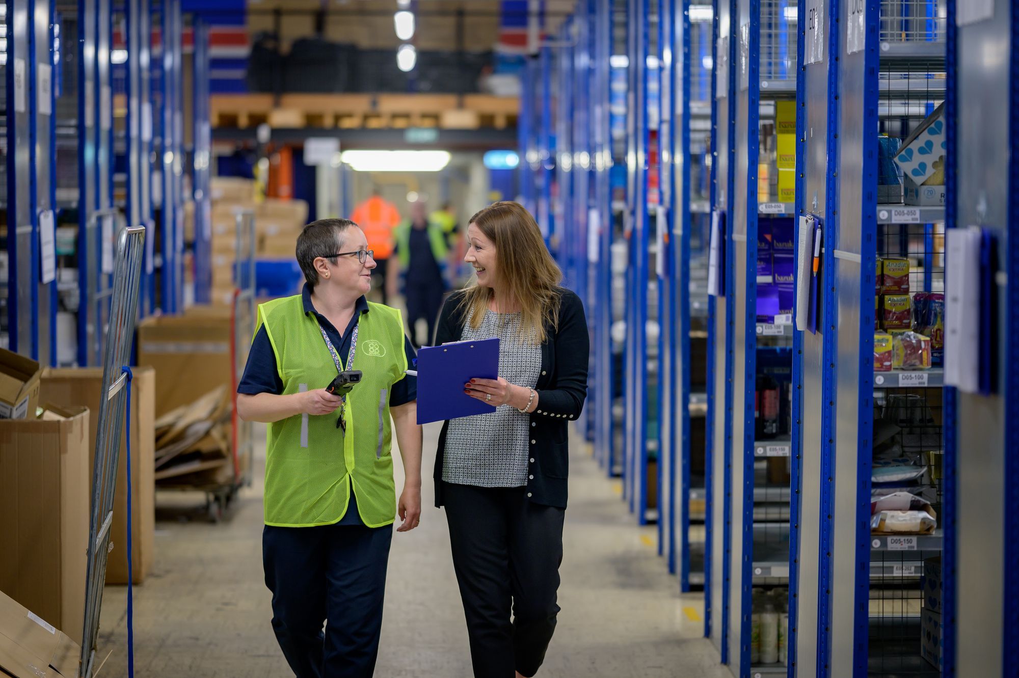 Two warehouse staff in hi-vis walking an aisle between storage racks
