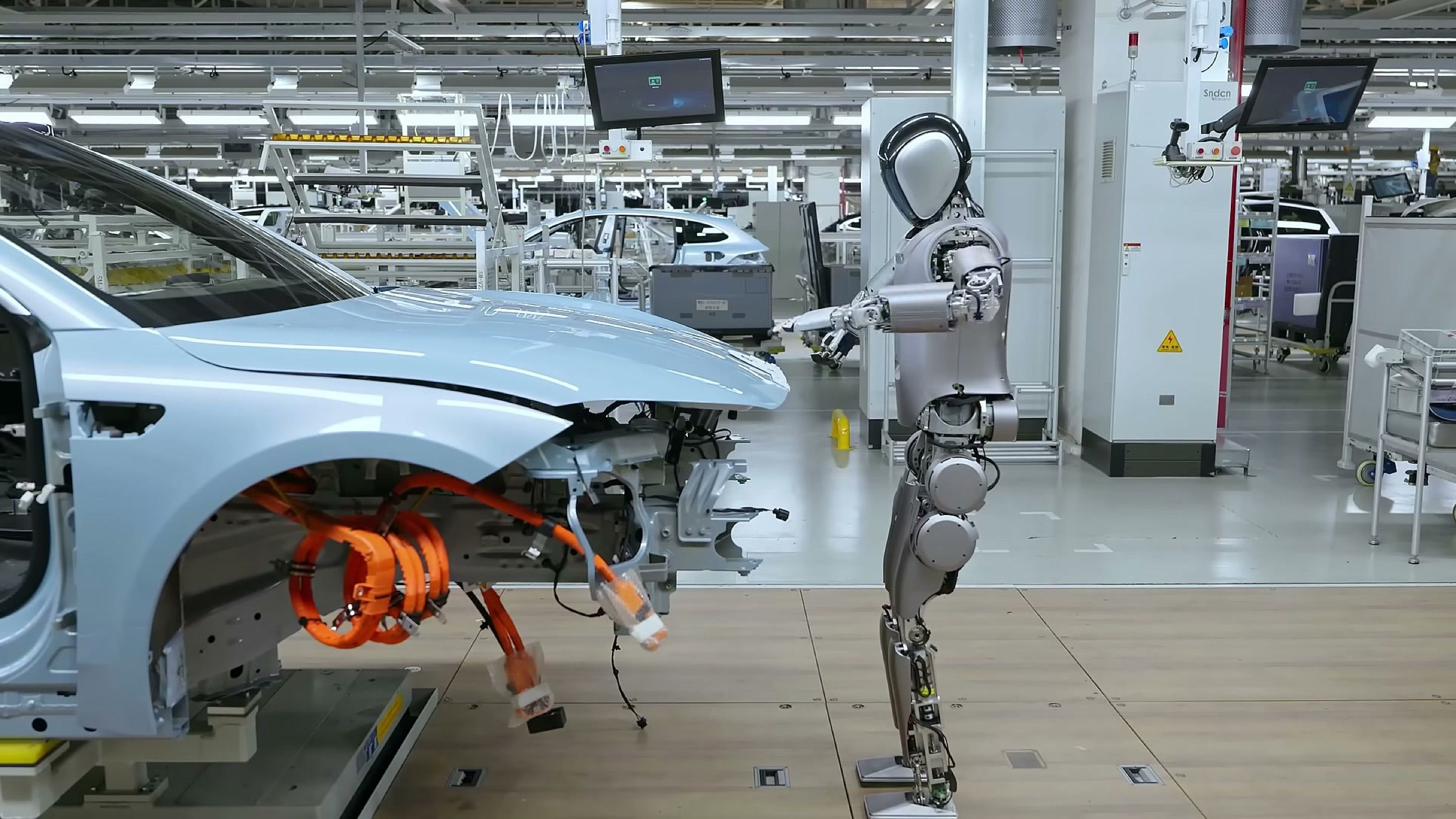 A humanoid robot stands at a workstation on an electric vehicle production line, checking a partly assembled car body.