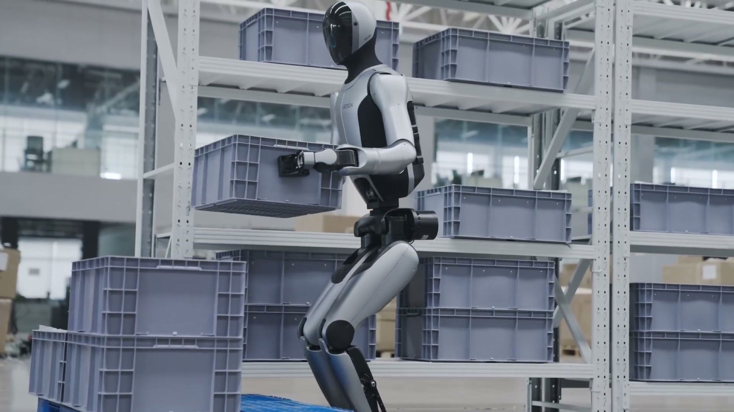 A humanoid robot lifts a plastic tote and places it onto a storage rack in a large warehouse.