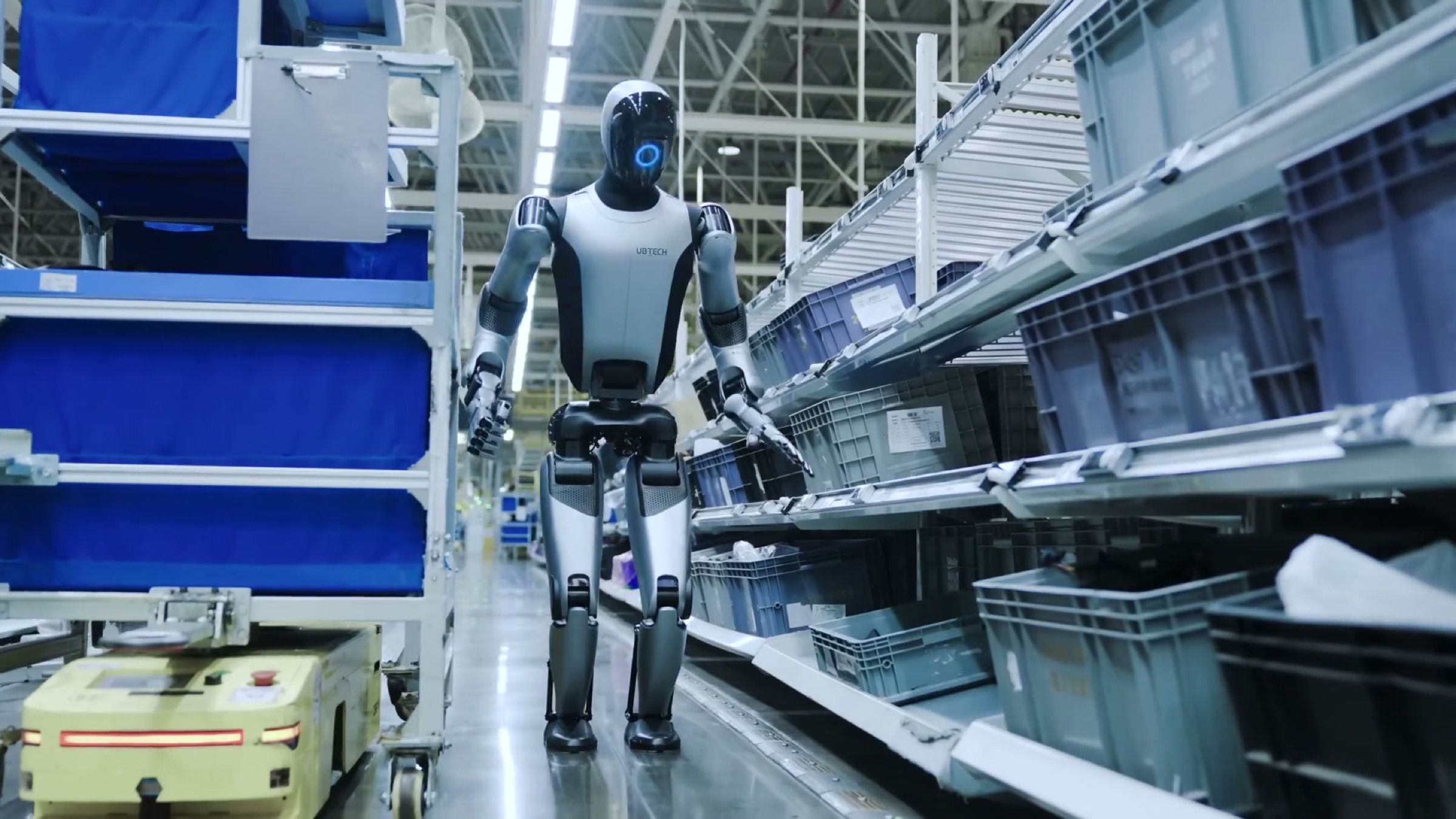 A humanoid robot works along a narrow parts aisle, reaching into shelved totes beside a mobile picking cart.