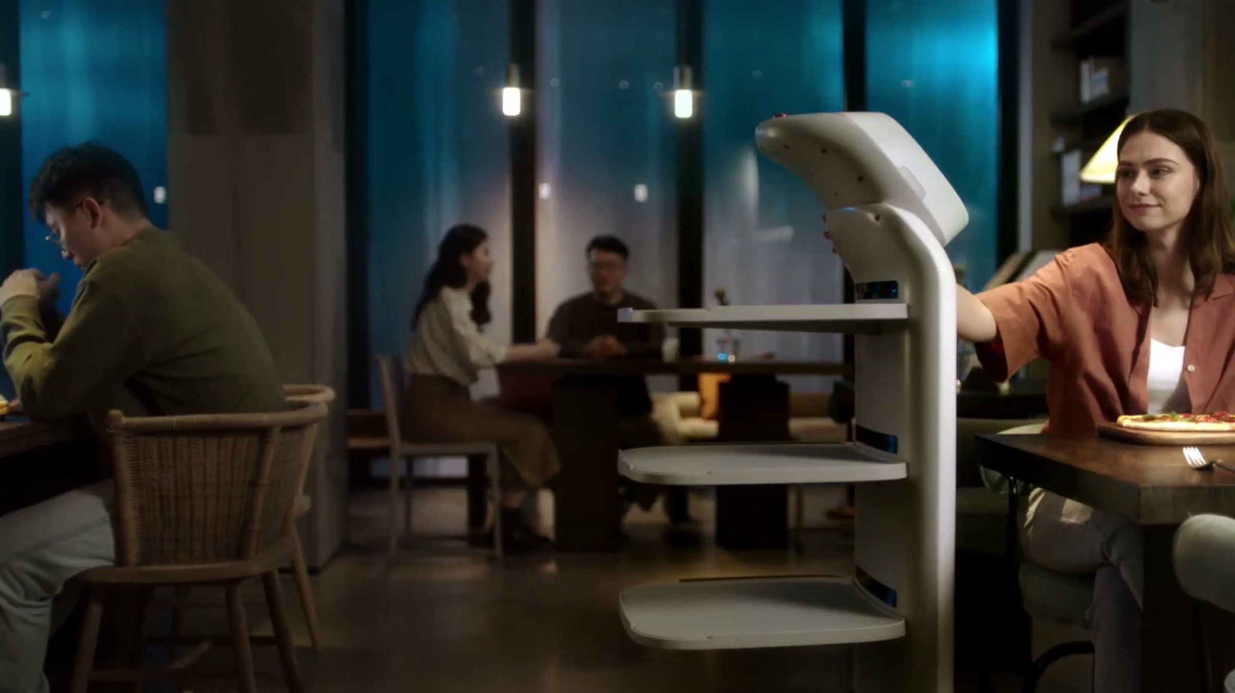 A tray-carrying delivery robot pulls up at a restaurant table as a diner reaches for her order.
